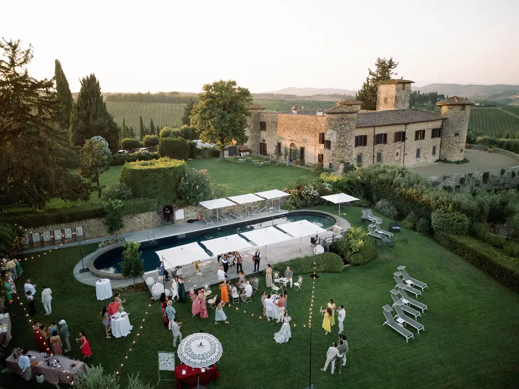 An outdoor wedding by a villa, featuring guests around a garden and pool, with string lights and a countryside backdrop.